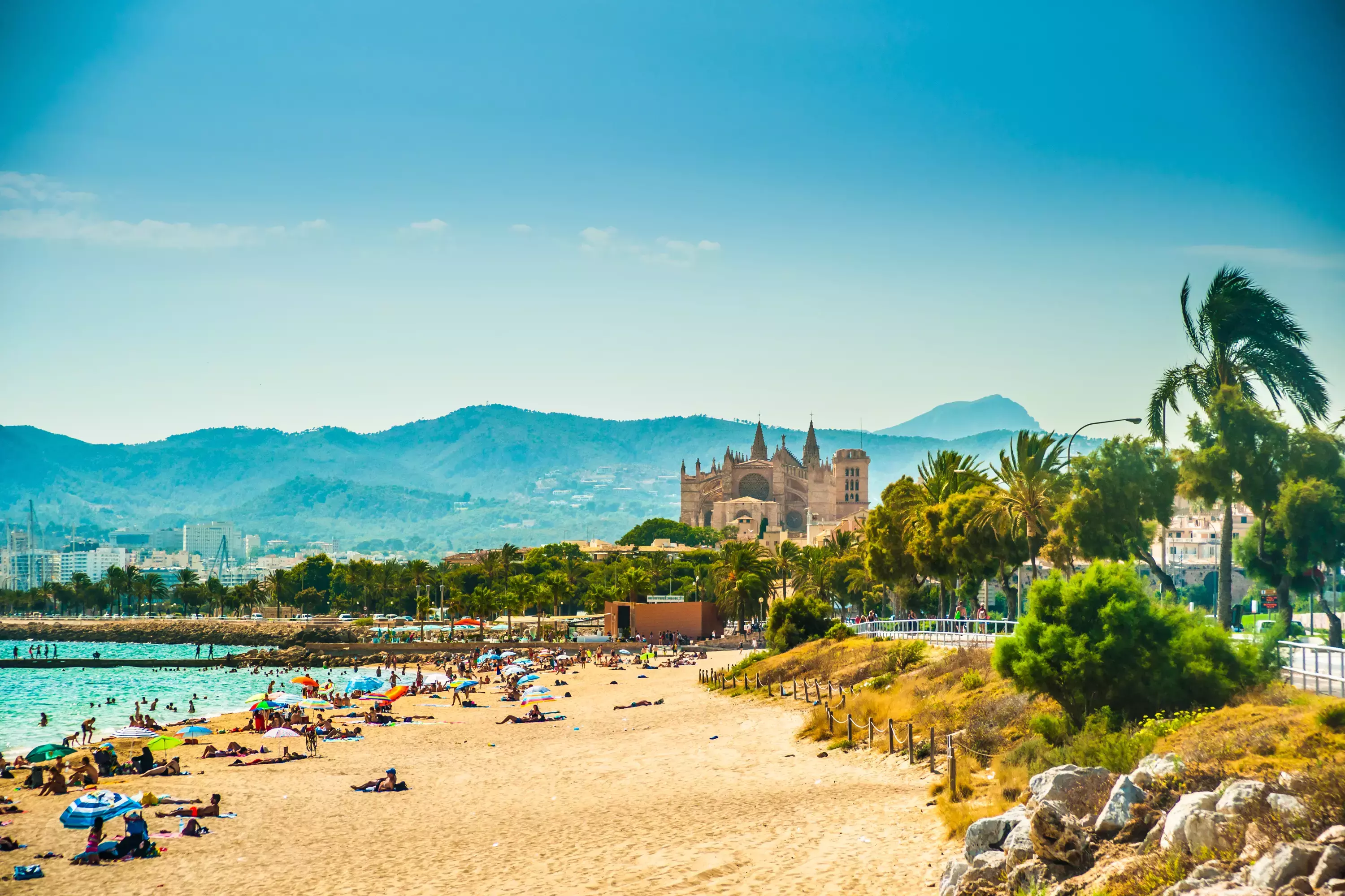 View of the beach of Palma de Mallorca with people lying on sand and the gorgeous cathedral building visible in background.